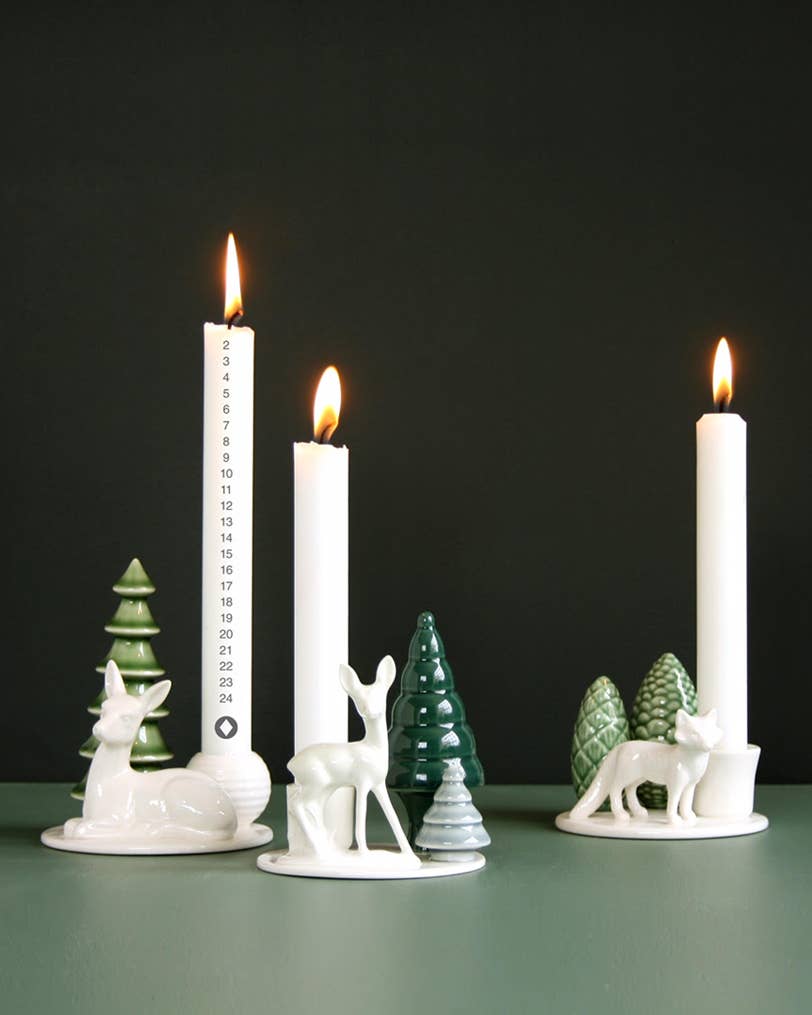 White porcelain deer, fawn, and fox candlestick holders with lit taper candles, a numbered advent candle, and ceramic evergreen trees in a Nordic winter table setting.