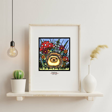 The Hedgehog Art Print by Sarah Angst, a framed giclée reproduction (11"x14" or 8"x10"), features a smiling hedgehog among mushrooms and flowers, displayed on a white shelf with woodland-themed decor accents.