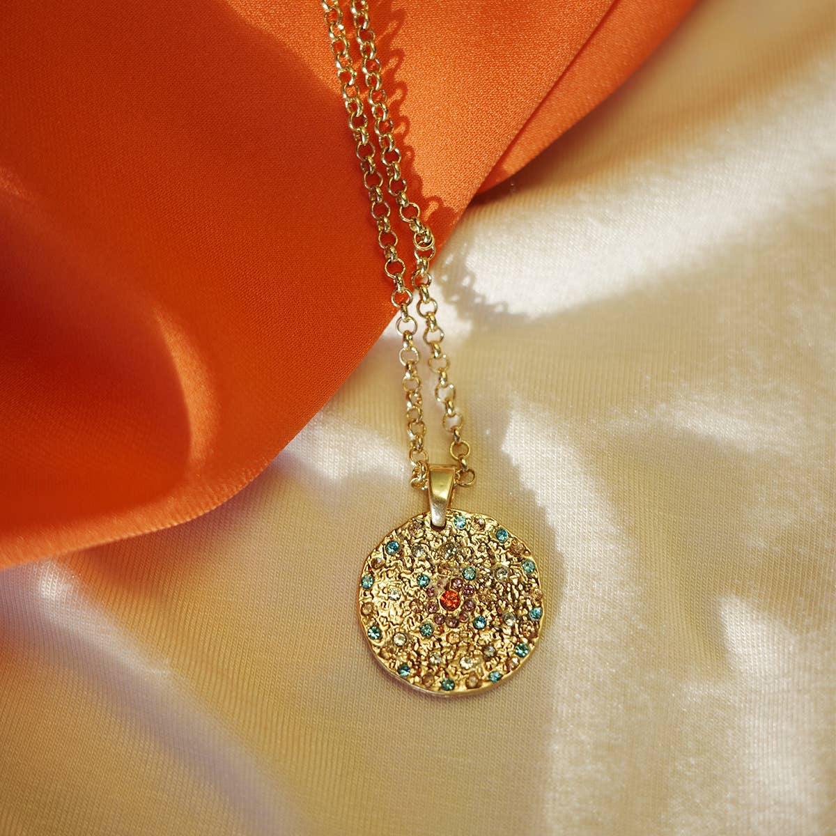 The A&C Oslo Vibrance Gold Coin Necklace features a textured pendant with colorful stones, ideal for layering. Shown on shiny cream fabric next to an orange cloth, it creates a vibrant display.
