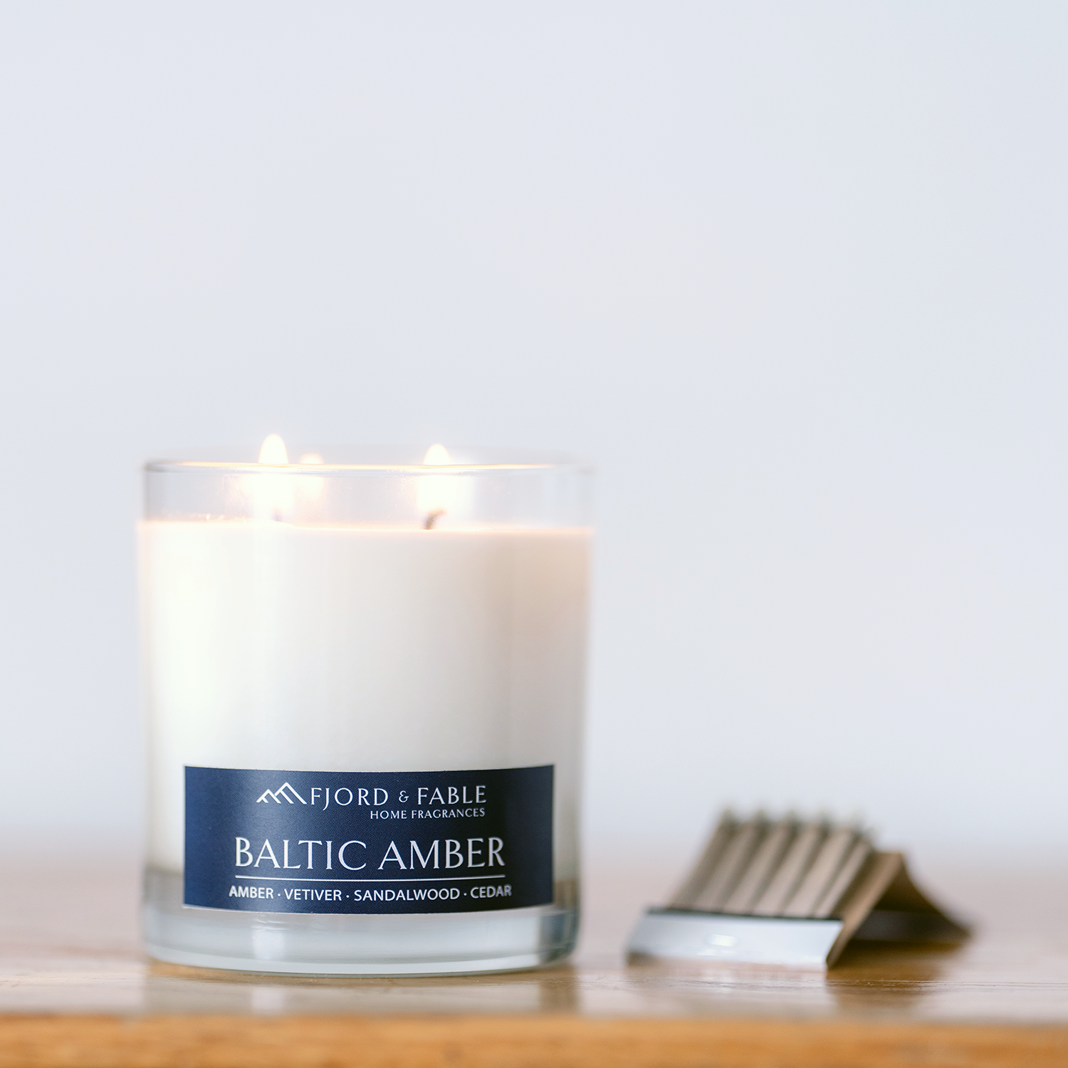 A glowing Fjord & Fable Baltic Amber Candle (4oz, amber, vetiver, sandalwood, cedar) sits in a glass container on a wooden surface beside a silver matchbox.