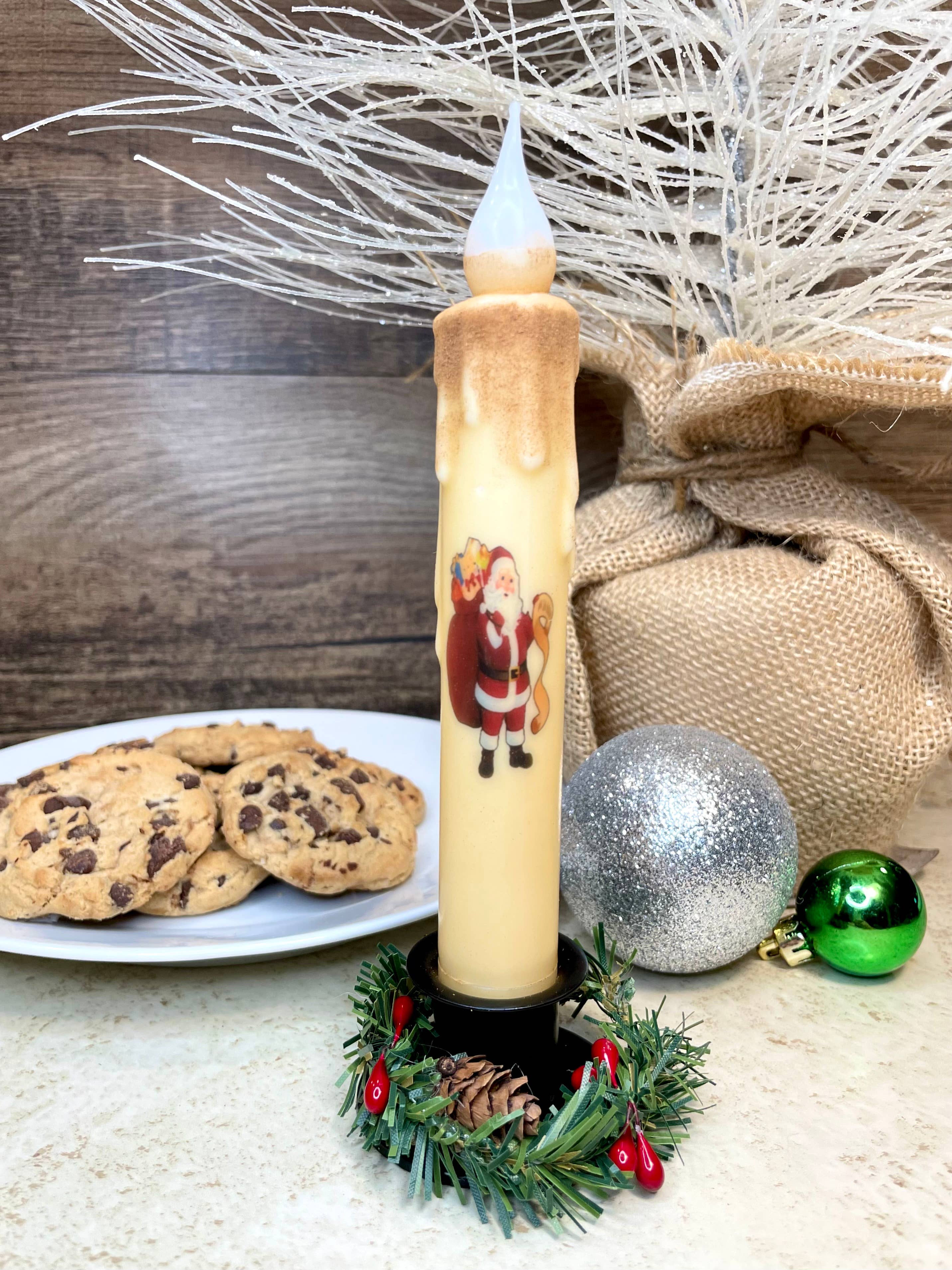 A Santa-shaped LED candle, featuring a battery-operated timer, stands on a decorated holder. Nearby are chocolate chip cookies on a plate, alongside silver and green ornaments and a burlap-wrapped item adorned with white branches.