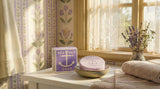 A bar of Sea Salt Lavender Swedish Dream Bar Soap – Triple Milled Natural Lavender 4 oz and its box rest in a bowl on a towel near a window with lace curtains, in a floral-wallpapered bathroom with lavender sprigs.