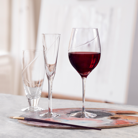 Three glasses—a mouth-blown tumbler, a KostaBoda: Line Flute Champagne Glass, and a wine glass with red wine—are artfully arranged on a painter’s palette next to a paintbrush.