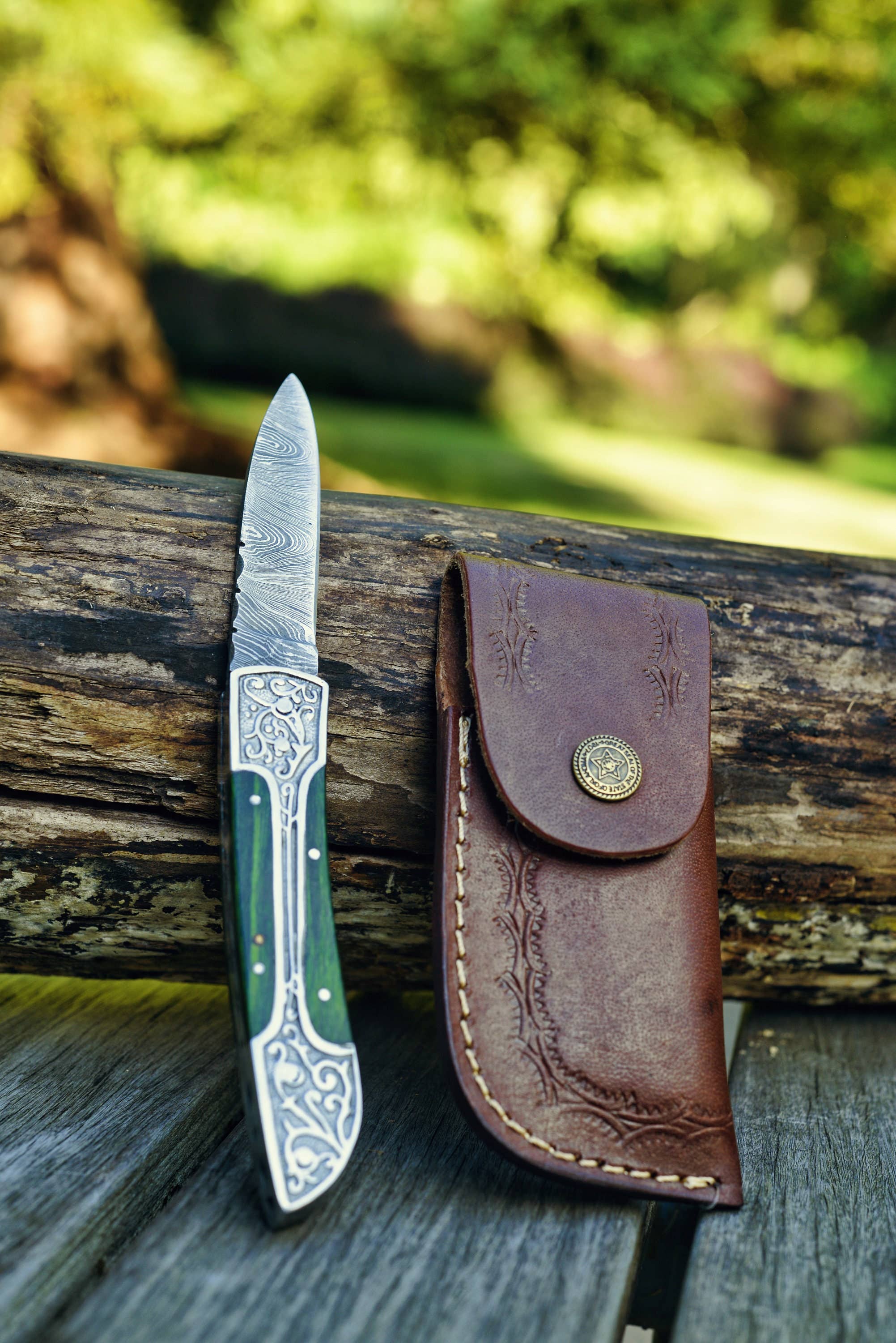 A Black Damascus Folding Knife Pocket Knife, featuring an intricate handle design, is elegantly placed beside a leather sheath on a rustic wooden surface with a softly blurred outdoor background. This genuine Damascus piece is perfect as a gift for men, seamlessly blending practicality with artistic craftsmanship.