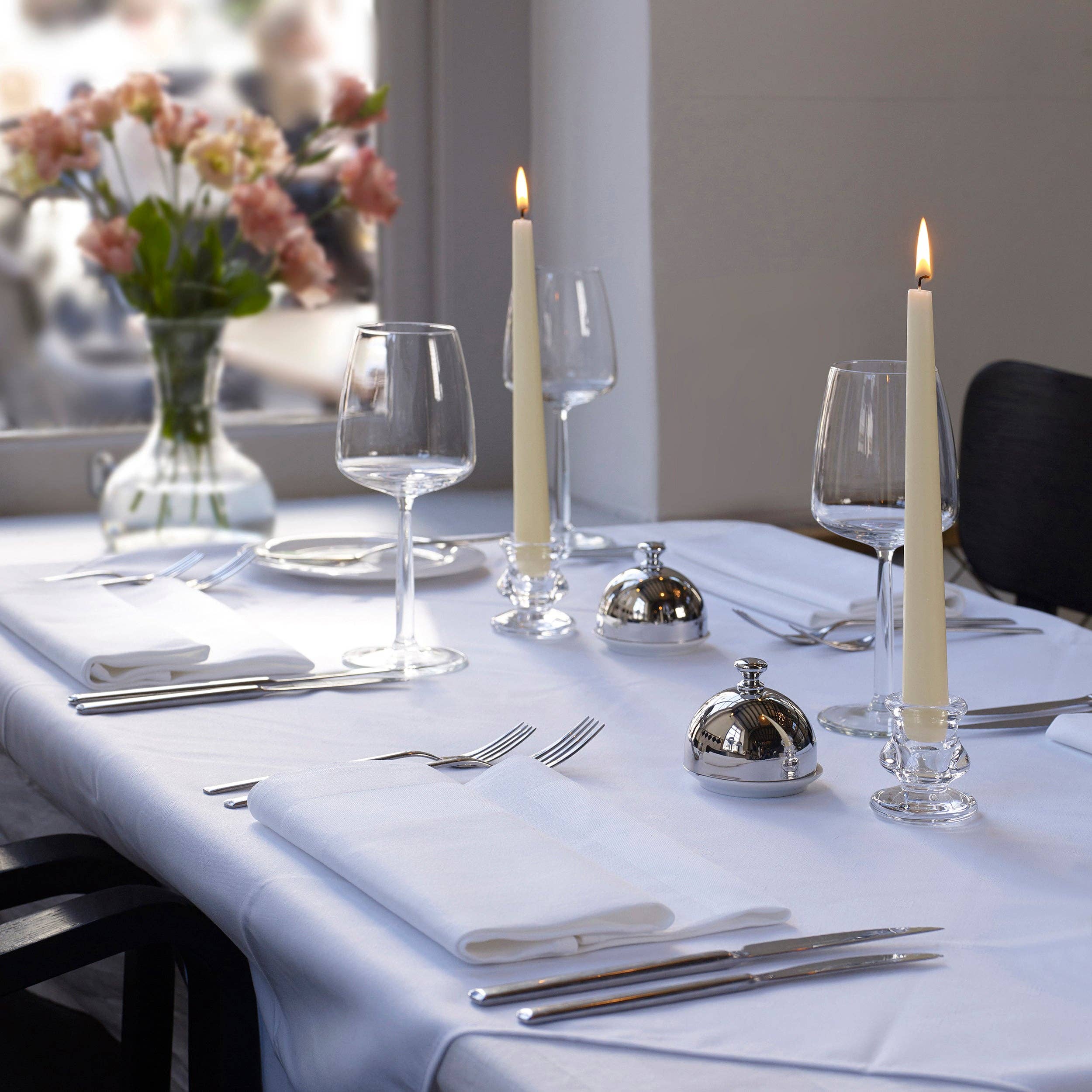 A dining table is set with white linens, two lit candles from the White Tall 10" Taper 4 Pack (Unscented), made from high-quality paraffin wax, alongside glassware, cutlery, and a vase of flowers by a window.