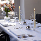 An elegant dining table setup includes a white tablecloth, warm glow from Tall 10 Inch Taper Candles, wine glasses, utensils, and a floral arrangement in a vase by the window.