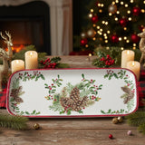 A tray with pinecone and holly design on a wooden table, surrounded by candles, greenery, ornaments, and a lit Christmas tree in the background.