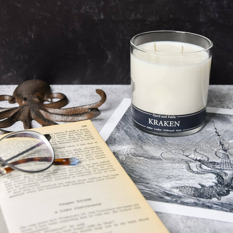 A Candle: Kraken - Amber, Sea Salt, Driftwood & Leather 8oz Jar by Fjord & Fable sits atop a black-and-white illustration beside an open book with glasses and an octopus figurine—ideal for lovers of mysterious scents.