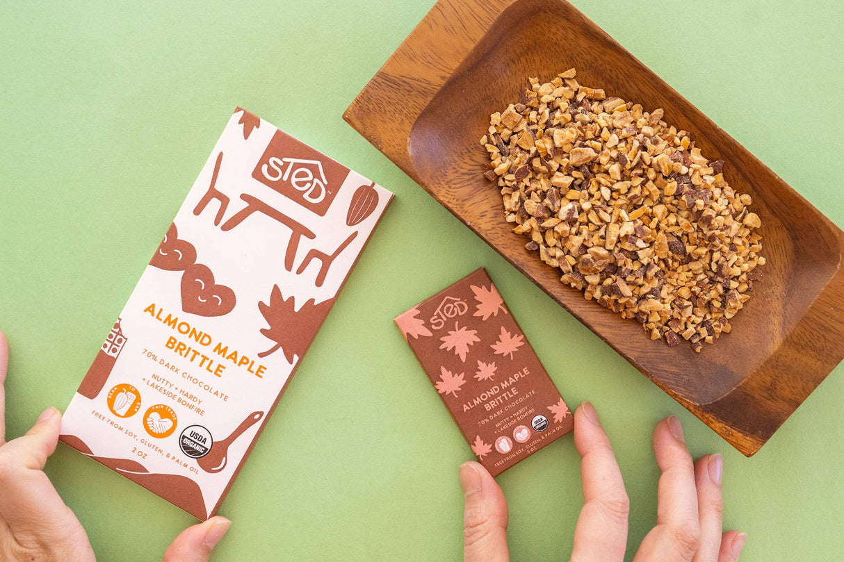 Hands hold almond maple brittle chocolate bars beside a wooden tray with brittle pieces on a light green surface.