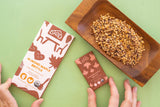 Hands hold almond maple brittle chocolate bars beside a wooden tray with brittle pieces on a light green surface.