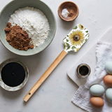 A spatula with a sunflower design on the handle lies on a countertop next to bowls of flour, brown sugar, eggs, a jar of spices, and a white towel.