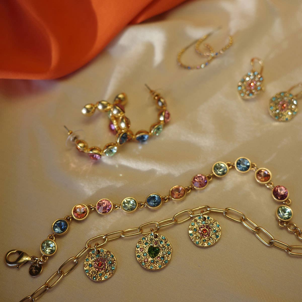 Vibrance Gold Hoop Earrings shine on a satin surface with gemstone bracelets, a necklace featuring round pendants, and an orange fabric partially visible in the corner.