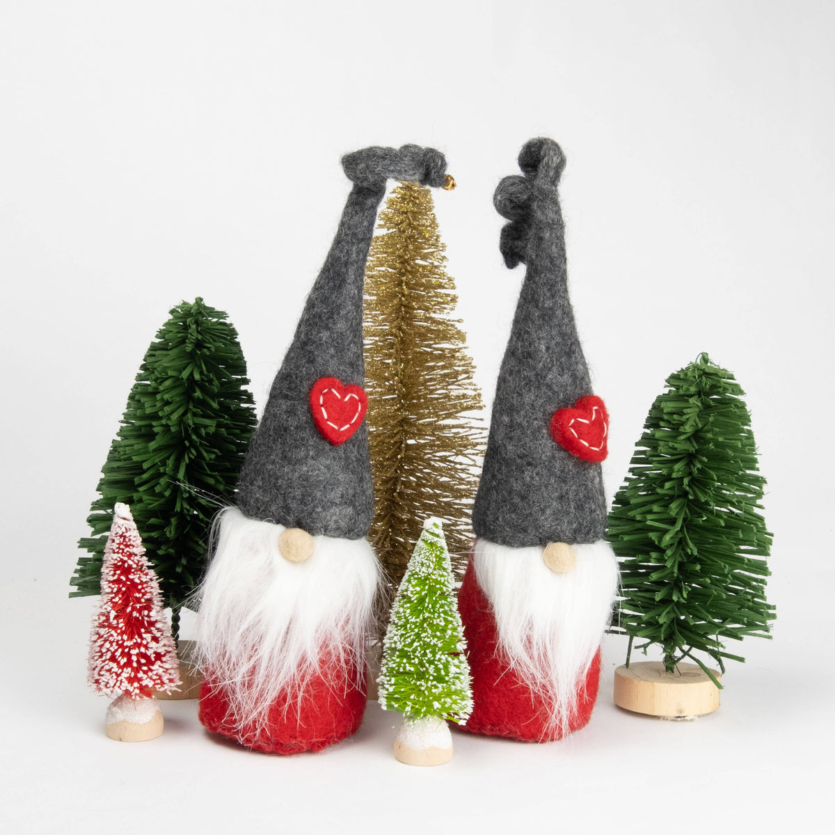 Two Scandinavian Tomte gnome figurines with tall hats and white beards stand among small artificial Christmas trees on a white background.