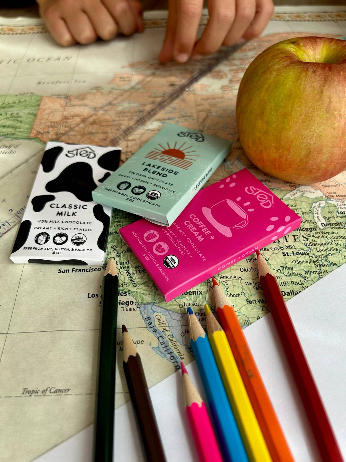 Three chocolate bars, including one labeled Ethiopian coffee chocolate, lie on a map near San Francisco with colored pencils and an apple; hands appear at the top.