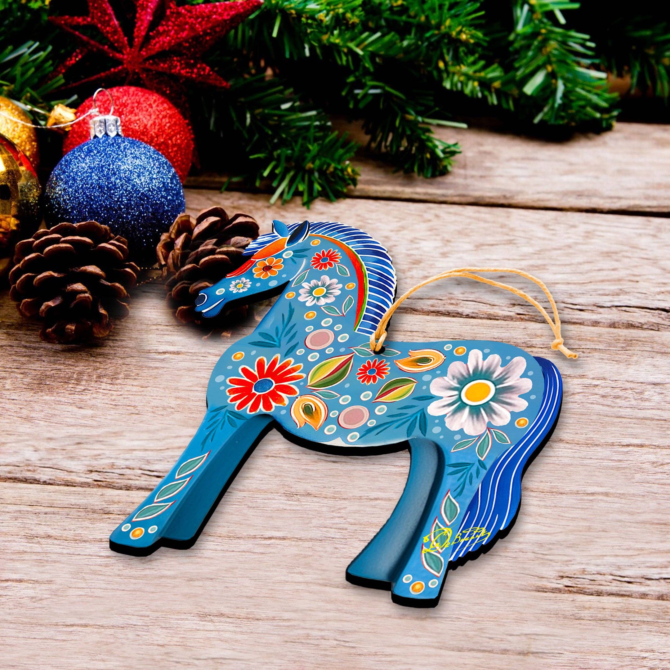 The Ornament: Danish Horse Decorative Wood Ornaments GDebrekht Love Family rests on a wooden surface, surrounded by pine cones, red and blue Christmas baubles, and green pine branches.