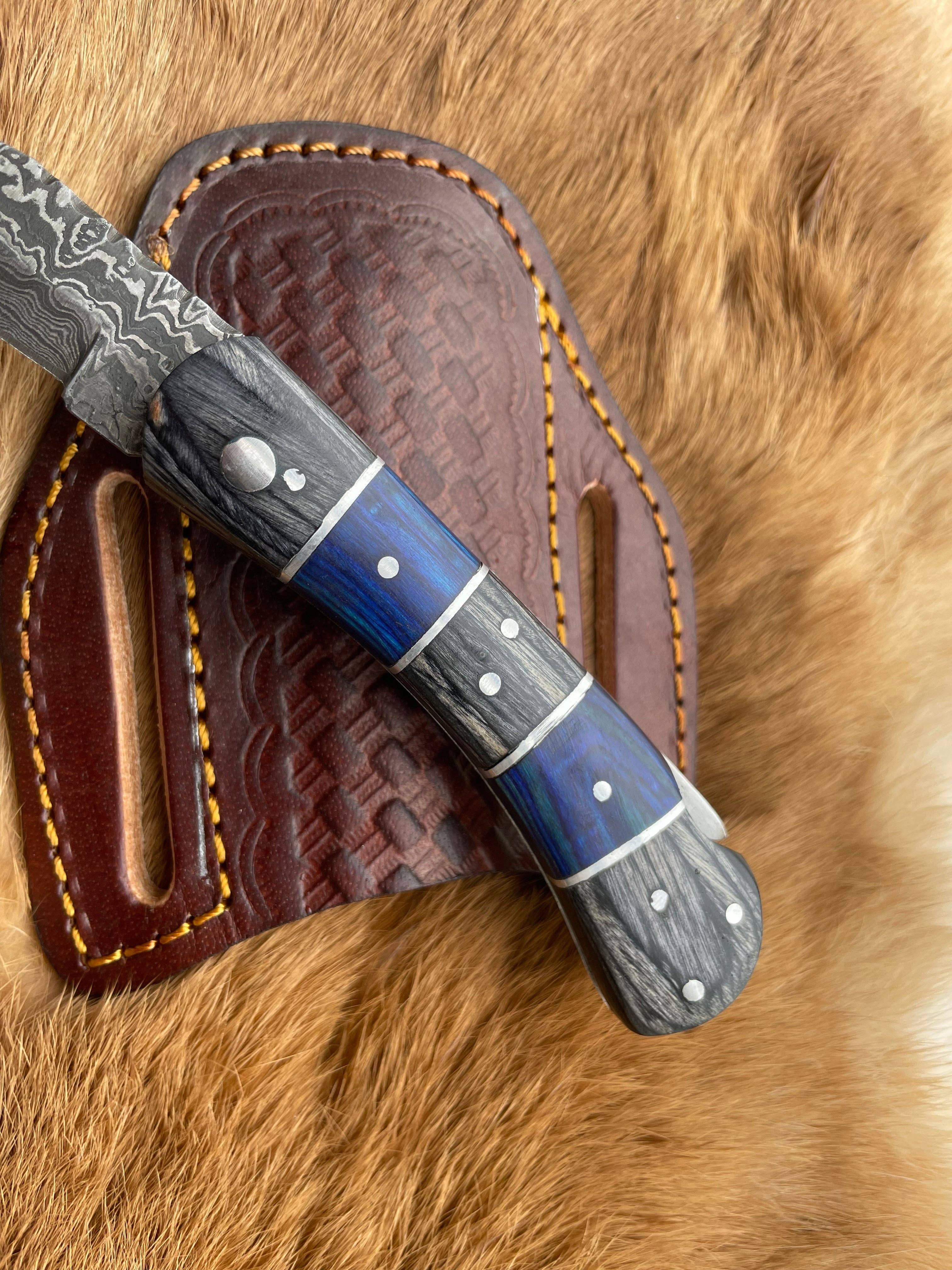 A close-up of the Knife: Grey and Blue Wood 6.5 Damascus Steel Folding Knife, featuring a patterned blade and blue-grey handle, resting on a brown leather sheath atop a fur surface.