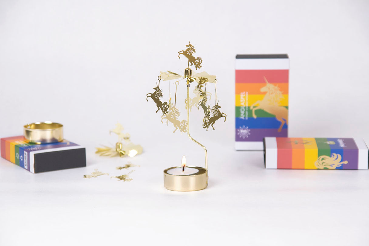 The Candleholder: Pluto Design Angel Chime Unicorn Rotary Candle Holder Gold adds a whimsical touch with rotating figures akin to Angel Chimes, placed next to vibrant packaging and a small tealight candle.