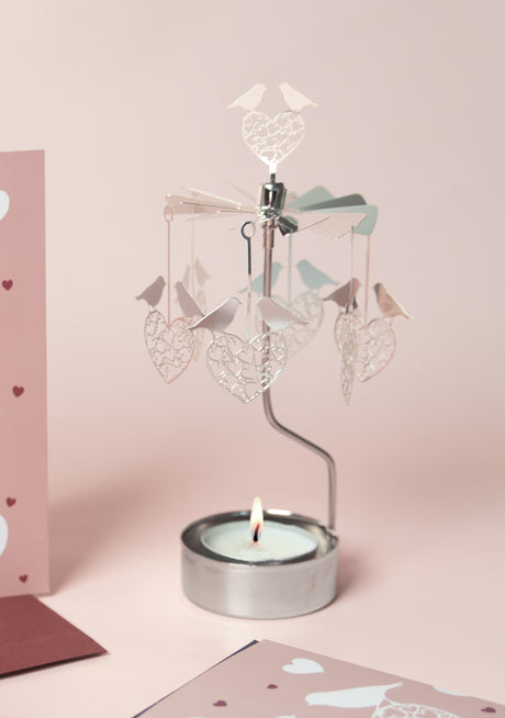 A lit tea light candle powers the Candle Holder: Love Birds Pluto Design Bird Heart "Love Birds" carousel, decorated with hanging bird heart shapes, set against a pink background with heart-patterned cards nearby.
