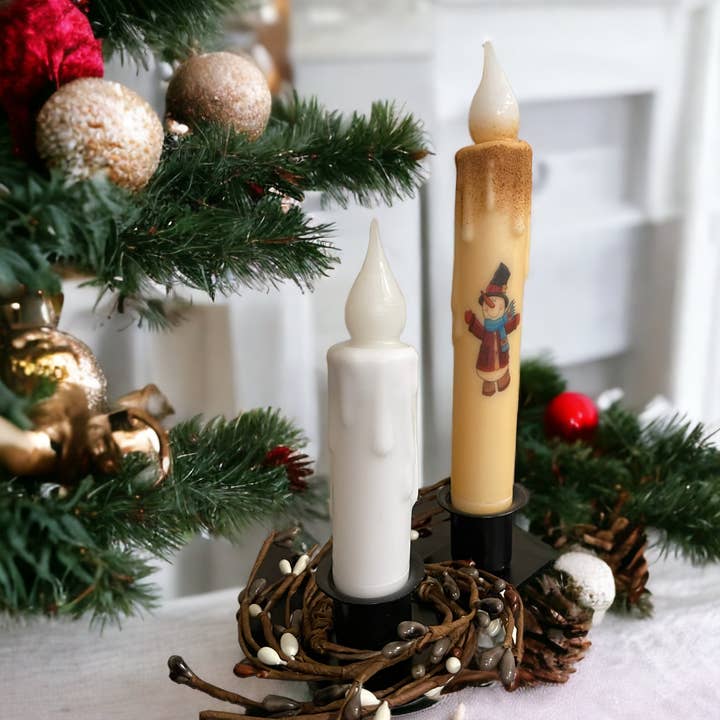 A pair of decorative candles features a solid white LED battery-operated timer taper candle, elegantly placed in a woven holder, accompanied by a snowman-designed candle. In the background, you can glimpse a Christmas tree adorned with ornaments.