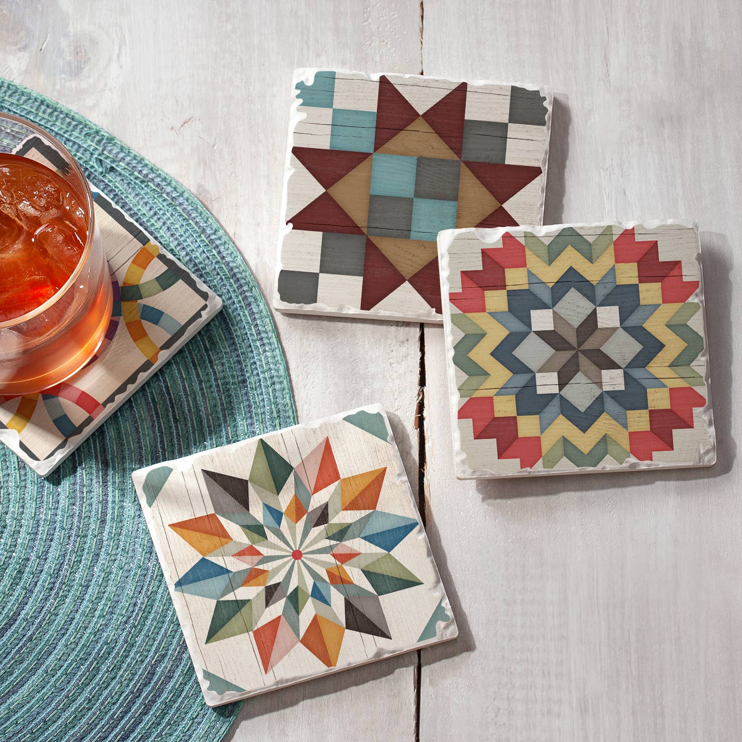 Three Barn Quilt Signs Stone Coasters with cork backing sit on a wooden surface, displaying vibrant geometric patterns. A drink rests on a green woven mat, showcasing their artisanal design from the 4-pack collection of coasters with four distinct images.