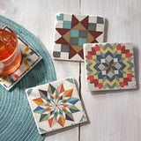 Four Coaster: Barn Quilt Signs Stone Coasters with Cork Backing (4 pack, 4" square) featuring colorful patterns are arranged on a white surface, one holding an iced drink. A round woven placemat adds Scandinavian decor charm.