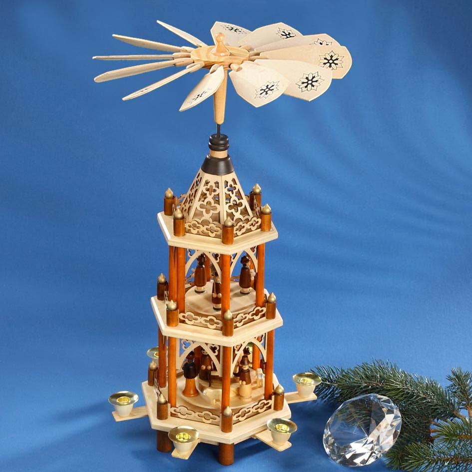 A beautifully crafted German 2-tier laser Christmas Pyramid stands gracefully on a blue background, its wooden blades spinning gently above candle holders. Next to it, a pine branch and crystal ornament add charm to this display of traditional craftsmanship.