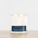 A glowing 4oz Baltic Amber Candle rests in a clear glass jar on light wood. Made with soy wax and essential oils, it fills the air with amber, vetiver, sandalwood, and cedar for a warm scent.