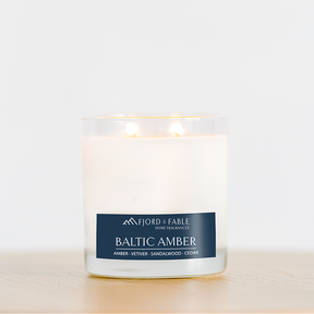 A glowing 4oz Baltic Amber Candle rests in a clear glass jar on light wood. Made with soy wax and essential oils, it fills the air with amber, vetiver, sandalwood, and cedar for a warm scent.