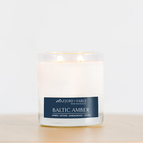 A glowing 4oz Baltic Amber Candle rests in a clear glass jar on light wood. Made with soy wax and essential oils, it fills the air with amber, vetiver, sandalwood, and cedar for a warm scent.