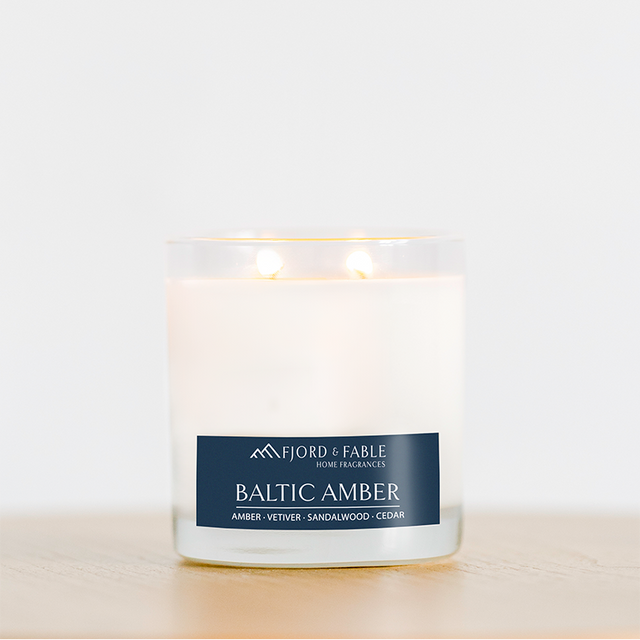 A glowing 4oz Baltic Amber Candle rests in a clear glass jar on light wood. Made with soy wax and essential oils, it fills the air with amber, vetiver, sandalwood, and cedar for a warm scent.