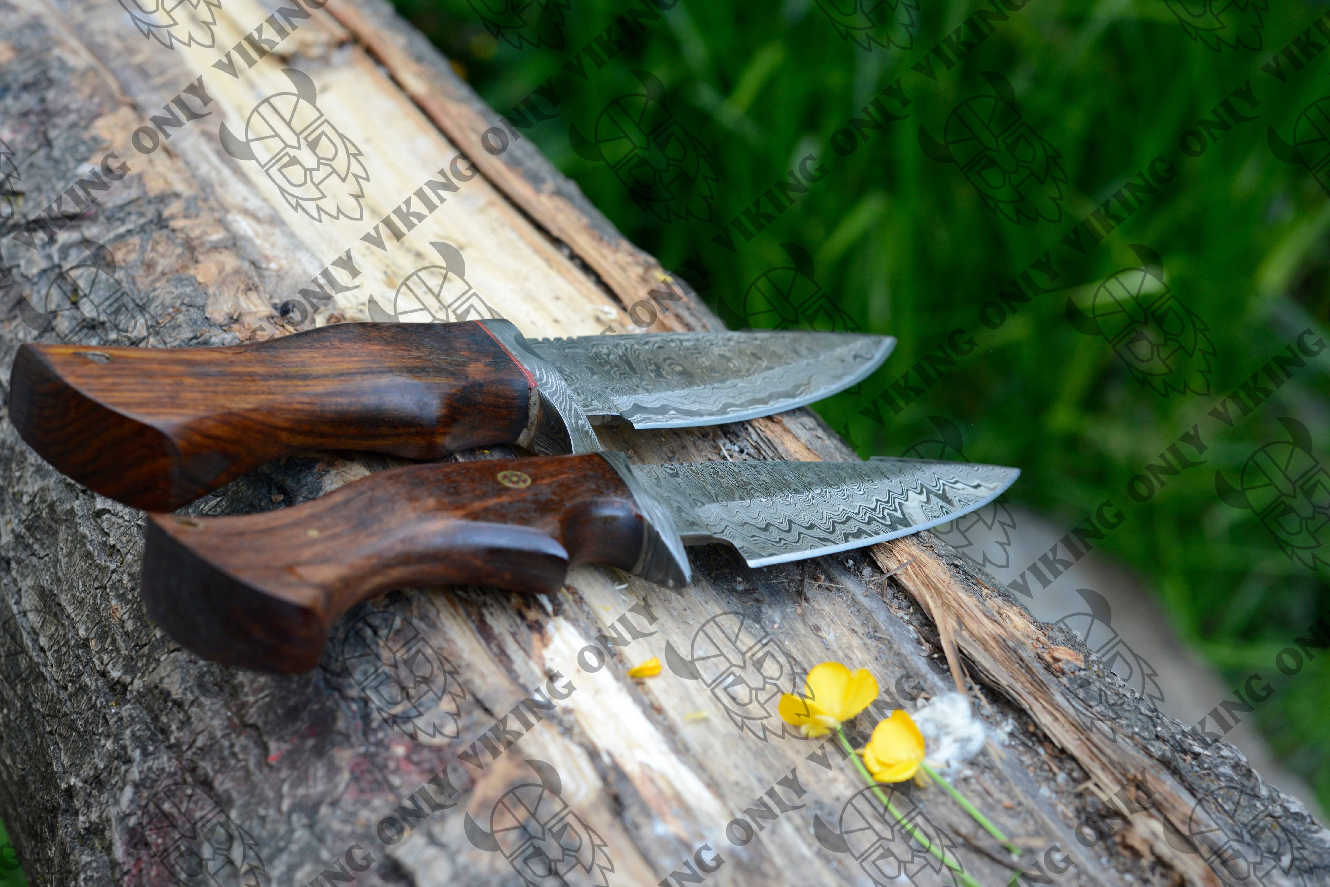 Two Premium Quality Utility Damascus Knives with wood handles and patterned blades rest on a tree trunk beside small yellow flowers. Ideal as camping tools, they blend seamlessly into the natural backdrop, with grass visible in the distance.