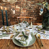 A rustic dining table features green and white plates, gold cutlery, pine napkins, candles, and festive decor arranged on the Pine Trees Wood Serving Tray (6x12”)—made in Sweden for Koustrup Co—set in front of an exposed brick wall.