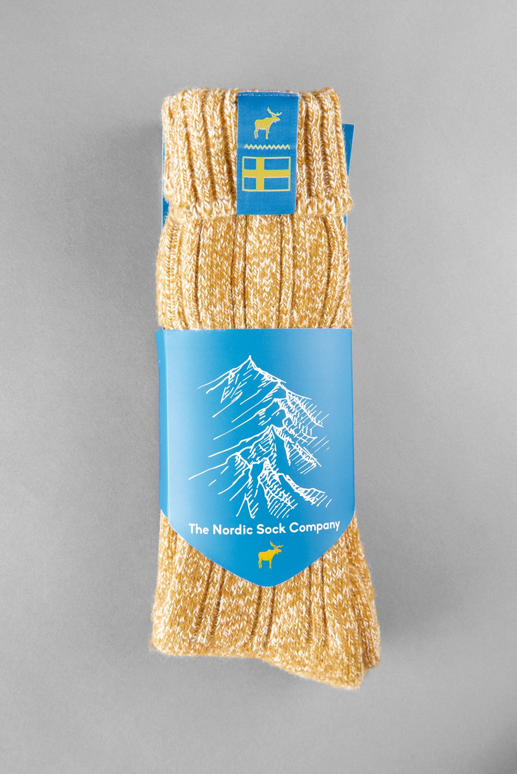 Swedish Lagom socks are knit in yellow and come with a blue packaging label showcasing a mountain design along with the text "The Nordic Sock Company." Made from a premium comfort wool blend, these unisex apparel essentials offer both warmth and style for everyone.