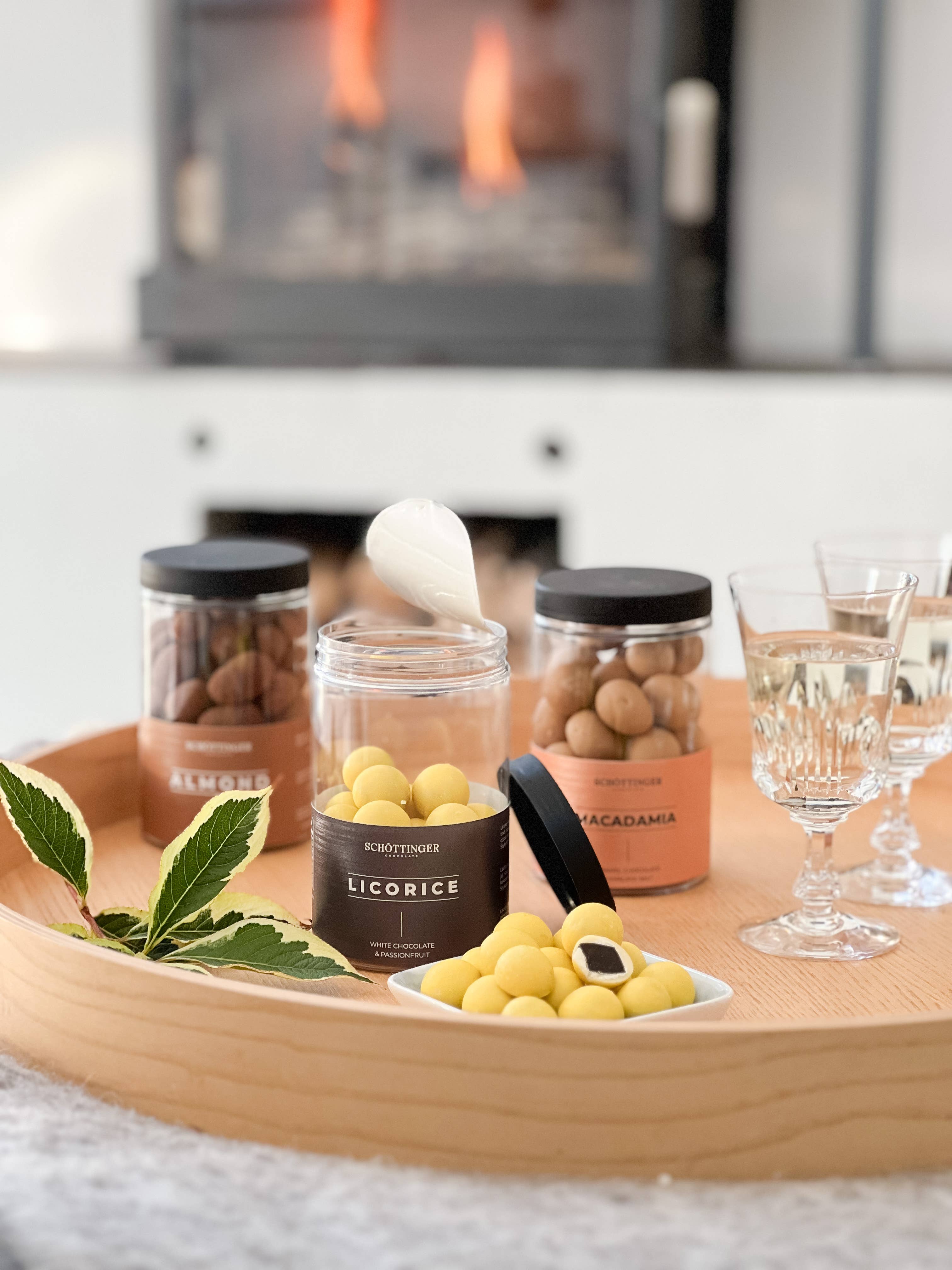 A wooden tray with Schottinger Licorice with White Chocolate & Passionfruit, almonds, macadamias, two glasses of water, and green leaves—set in front of a fireplace.