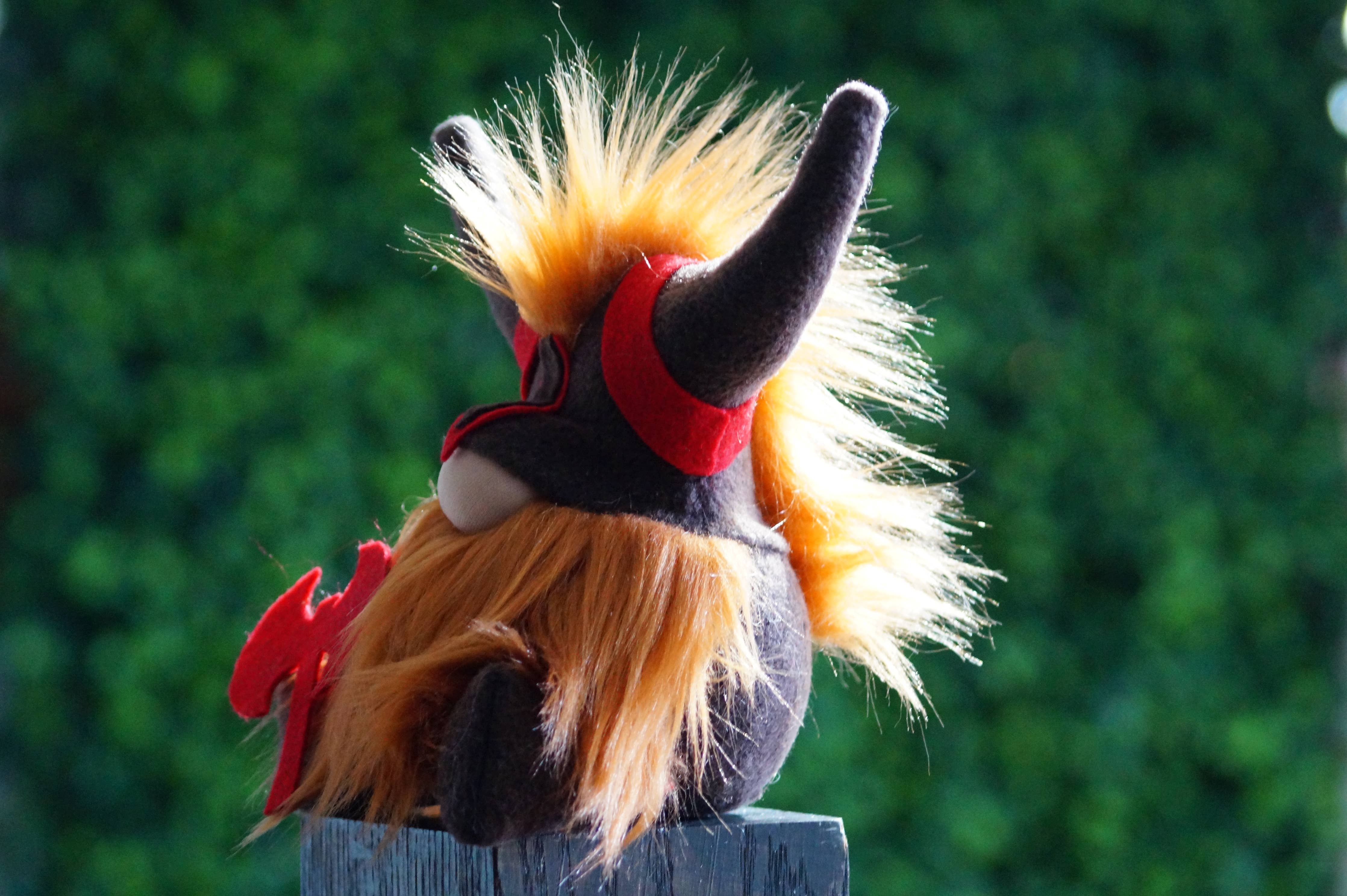 A Viking Gnome plush toy with long orange fur, a black body, a red mask, and large black horns sits on a wooden surface against a blurred green background. This unique gift captures the essence of whimsy and adventure.