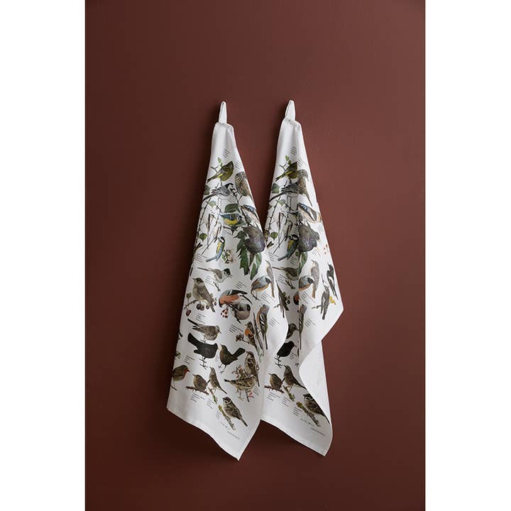 Two 100% organic cotton Garden Birds tea towels (19x27) by Carl Christian Tofte for Koustrupco hang on hooks against a brown wall.