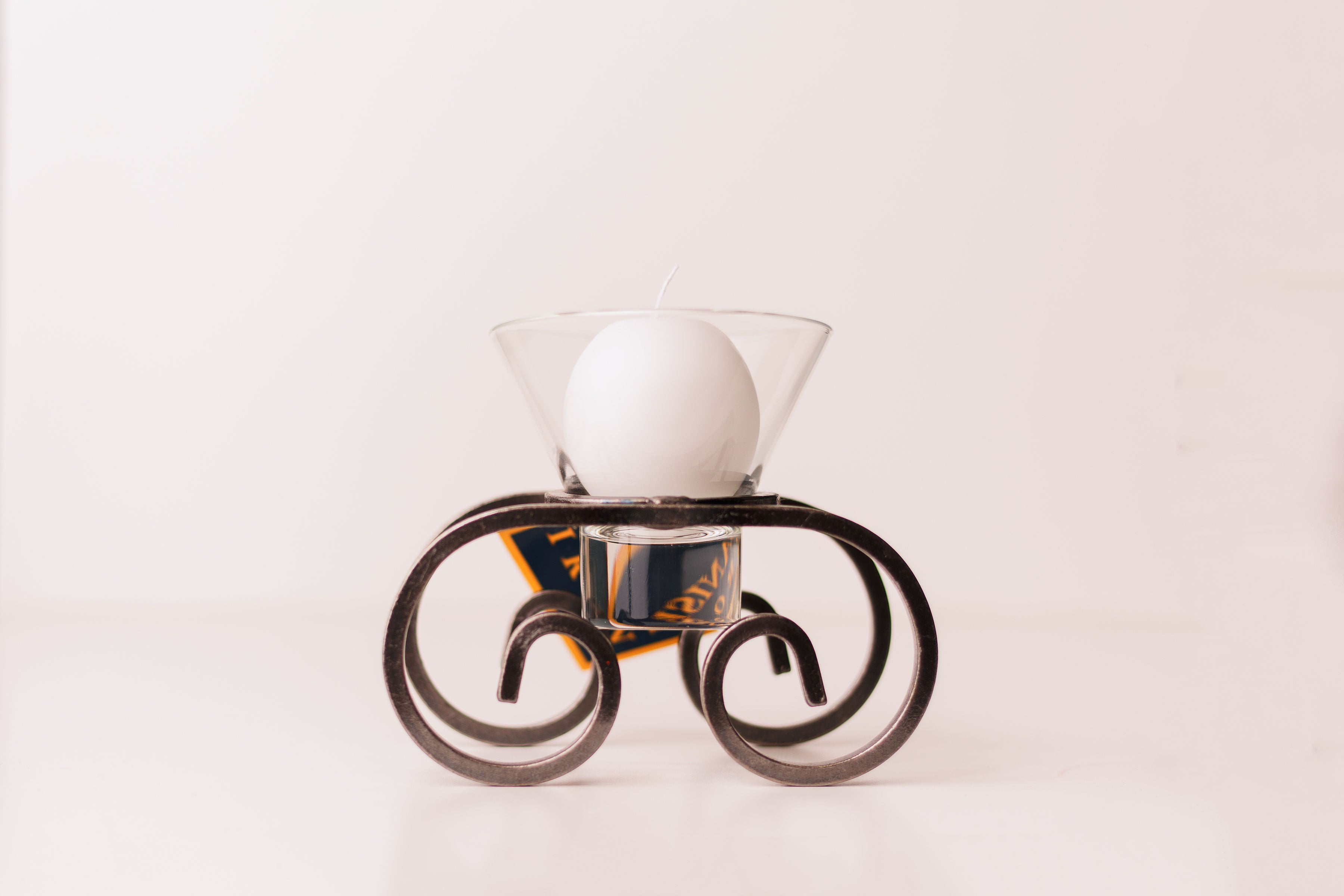 A white round candle in a glass cup sits on the Candle Holder: Danish Iron with Leif Glass Cup Ball Candle Holder, featuring curled legs, against a plain background.