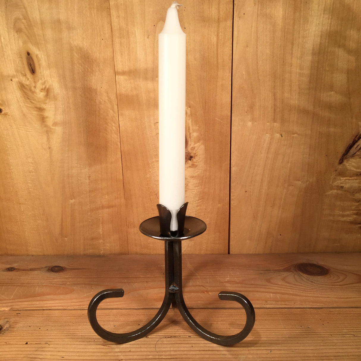 The "Danish Iron Taper Candleholder with Curly Bottom" elegantly displays a white candle on a wooden surface, with its curved base harmonizing beautifully against the backdrop of a wooden wall, enhancing the timeless charm of tapered candles.