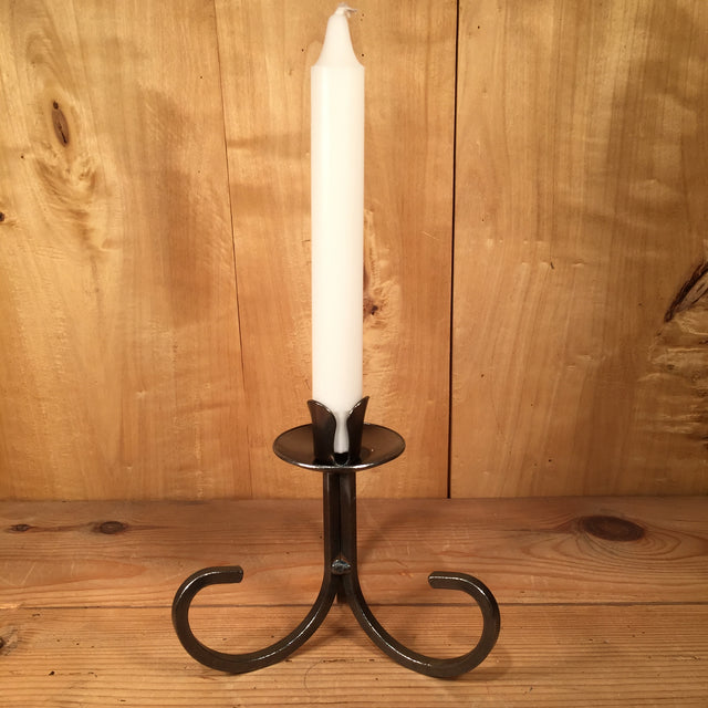 The "Danish Iron Taper Candleholder with Curly Bottom" elegantly displays a white candle on a wooden surface, with its curved base harmonizing beautifully against the backdrop of a wooden wall, enhancing the timeless charm of tapered candles.