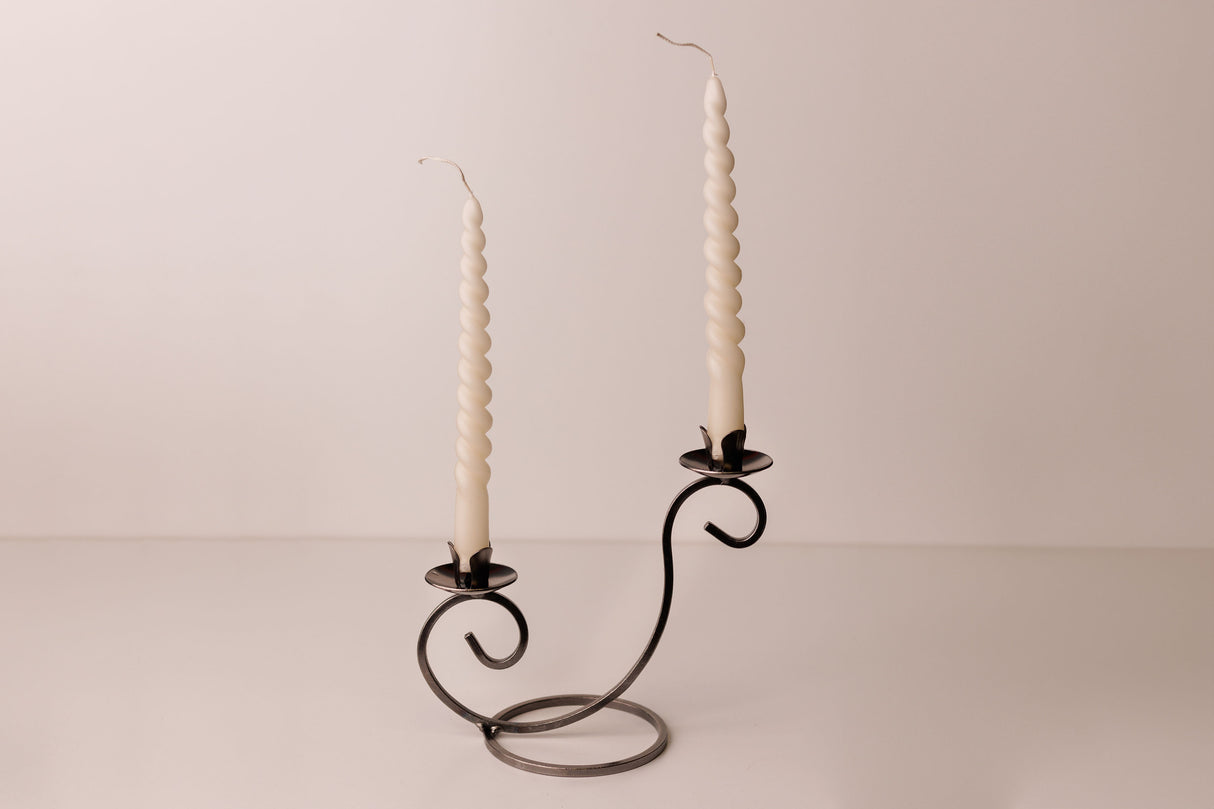 Two white spiral candles in a Danish iron holder with curved arms, set on a plain light surface with a neutral backdrop.
