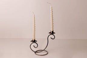Two white spiral candles in a Danish iron holder with curved arms, set on a plain light surface with a neutral backdrop.