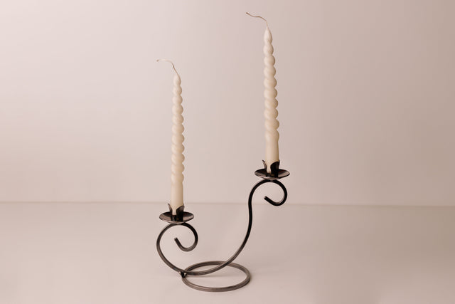 Two white spiral candles in a Danish iron holder with curved arms, set on a plain light surface with a neutral backdrop.