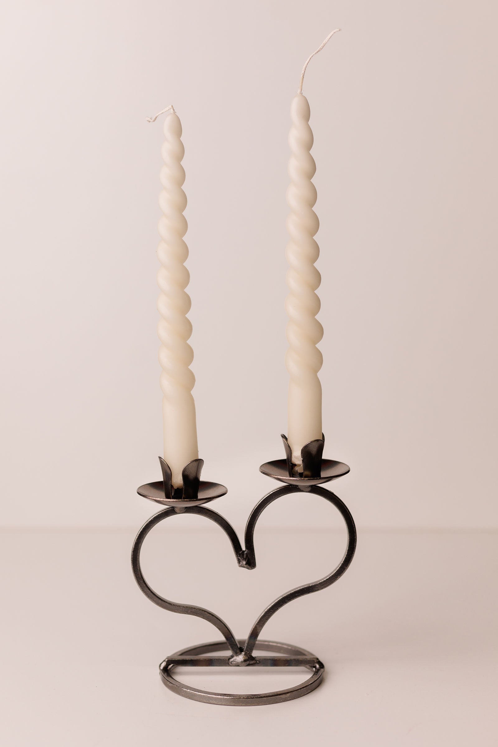 Two twisted white candles rest elegantly in a Danish Iron Taper Candleholder, featuring a large heart design that holds two taper candles, on a plain surface.