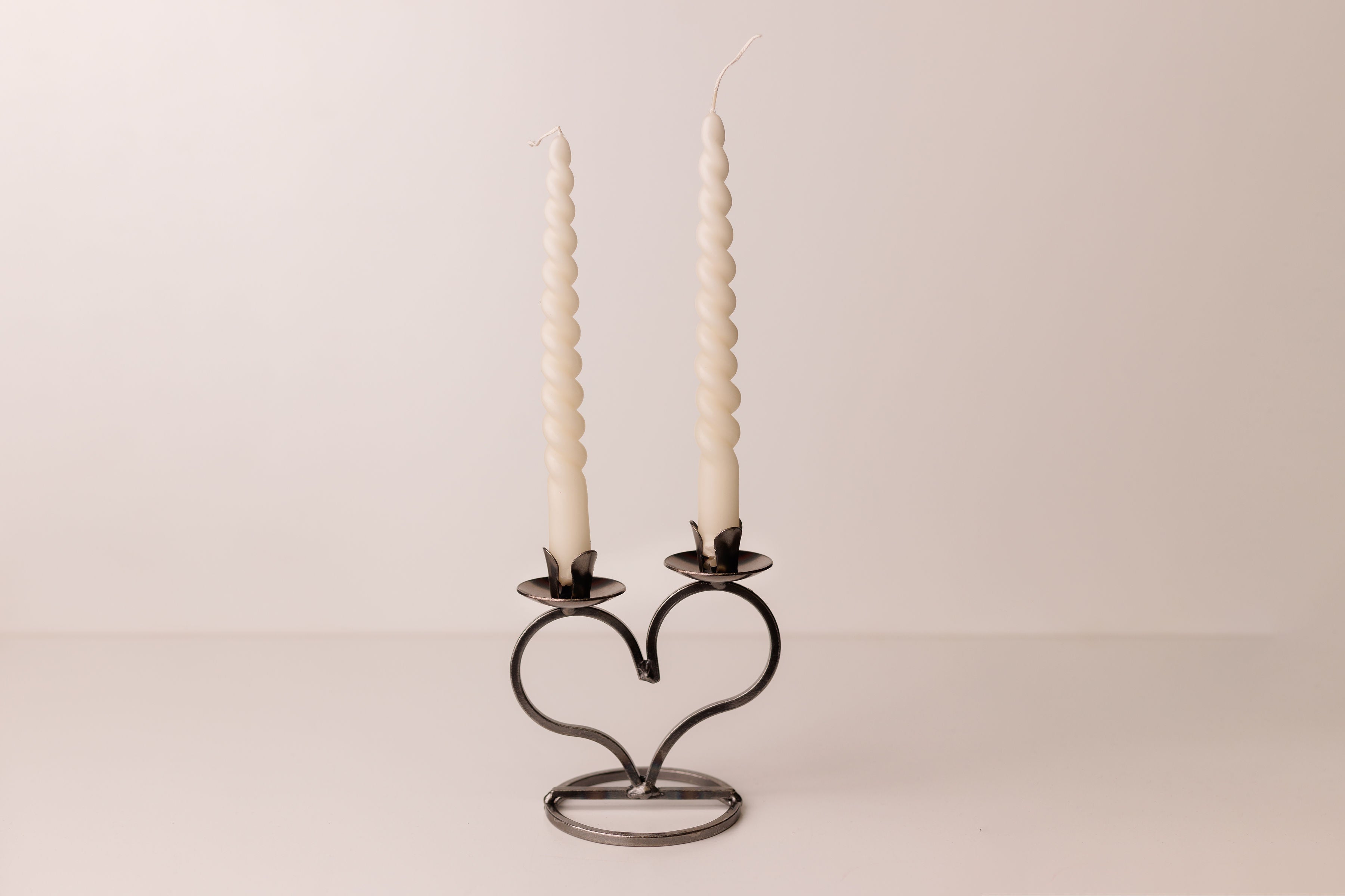 A pair of white taper candles sits gracefully in a heart-shaped Danish iron taper candleholder with a curly bottom, set against a simple background.