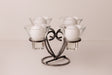Danish iron candle holder with four round white candles in clear glass cups, symmetrically arranged on a light surface.