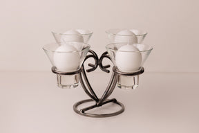 Danish iron candle holder with four round white candles in clear glass cups, symmetrically arranged on a light surface.
