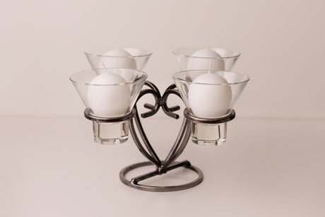 Danish iron candle holder with four round white candles in clear glass cups, symmetrically arranged on a light surface.
