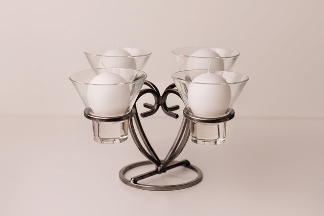 Danish iron candle holder with four round white candles in clear glass cups, symmetrically arranged on a light surface.