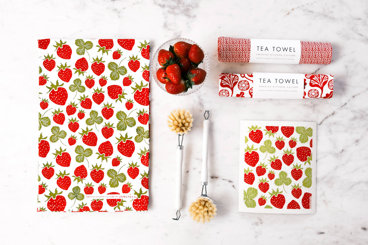 Flat lay of two tea towels with strawberry and red patterns, a bowl of strawberries, two dish brushes, and the Strawberries Swedish Dish Cloth on a white marble surface.