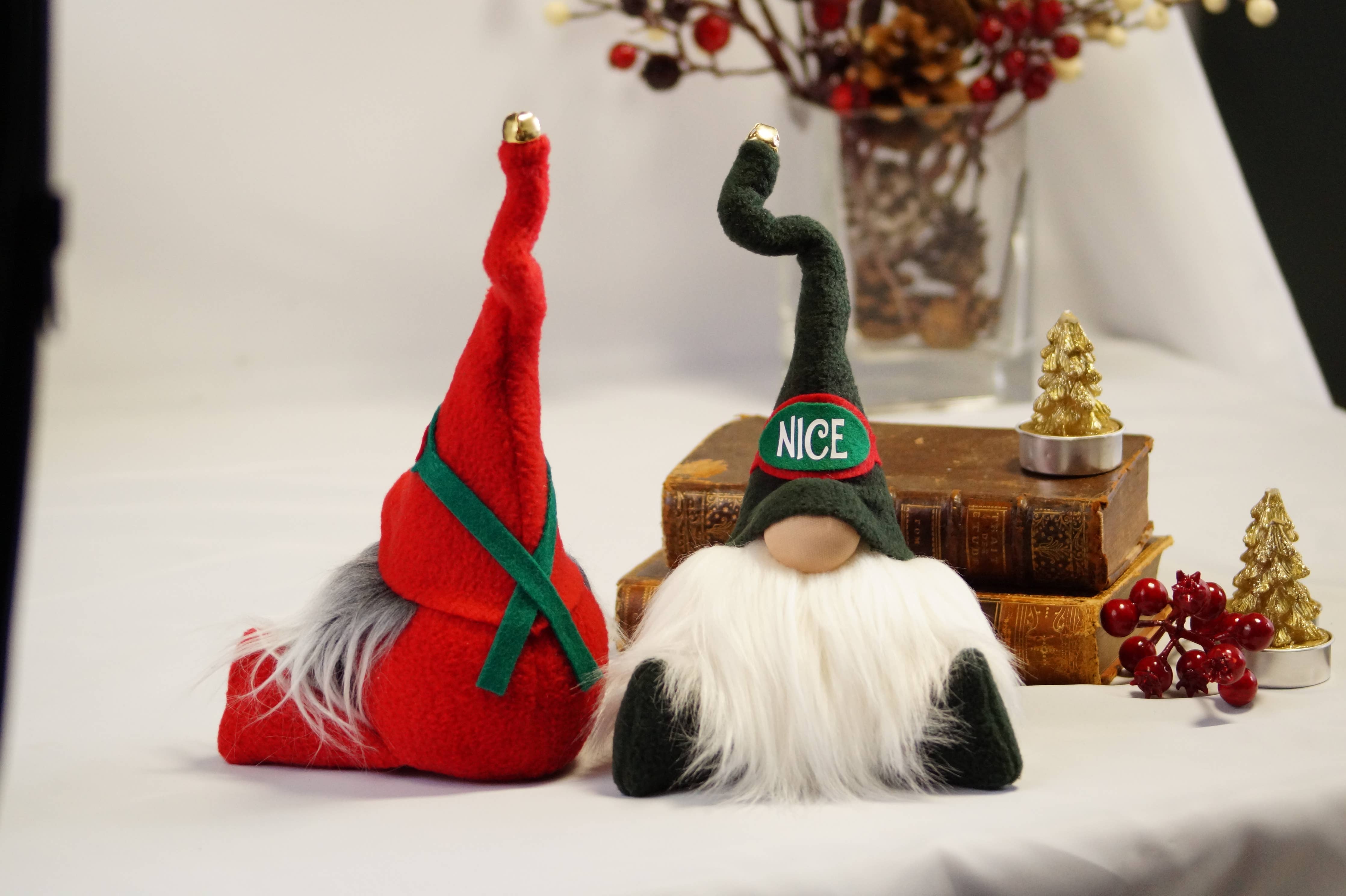 Two festive gnome figurines sit on a table, one wearing a green hat labeled "NICE" and the Naughty Christmas Gnome sporting a red one. They are surrounded by small holiday decorations, books, and hints of holiday games that promise laughter and cheer.